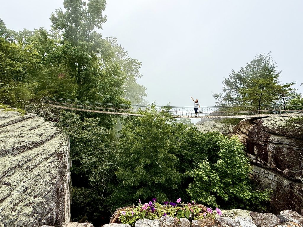 Woman standing on the swing-a-long bridge at Rock City overlooking a lush forest and rocky cliffs near Chattanooga, Tennessee
