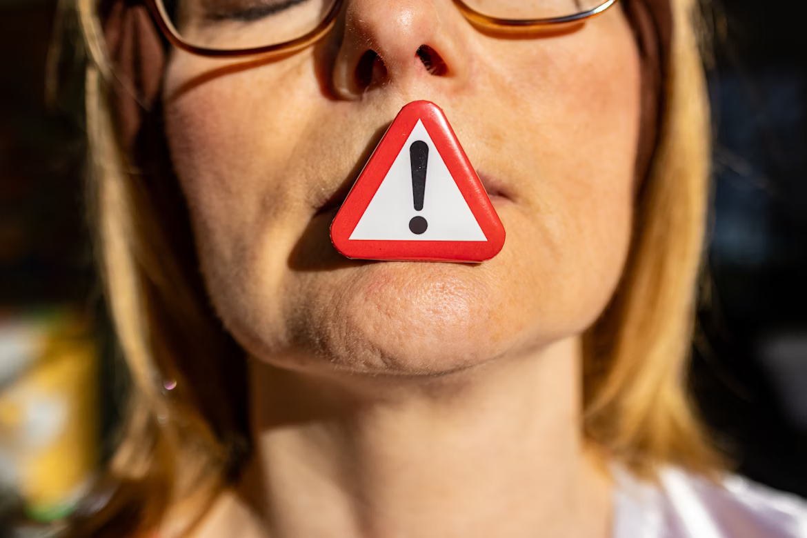 Person wearing glasses with a red warning triangle covering their mouth.