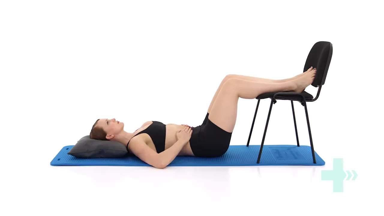 Person lying on back with hips and knees at 90 degrees on a chair, hands gently on the abdomen showing proper diaphragmatic breathing setup.