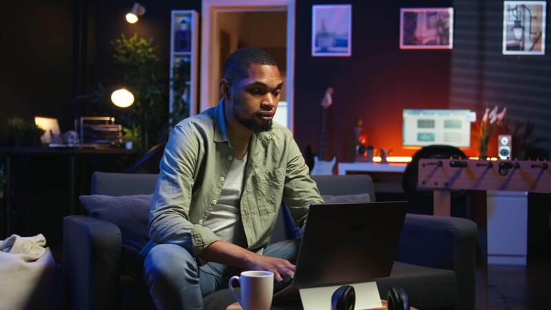 Young black guy working on his freelance tasks in the living room,  adjusting his job schedule and enjoying work from his cozy home. Person  navigating on web with laptop, multitasking. Camera A.