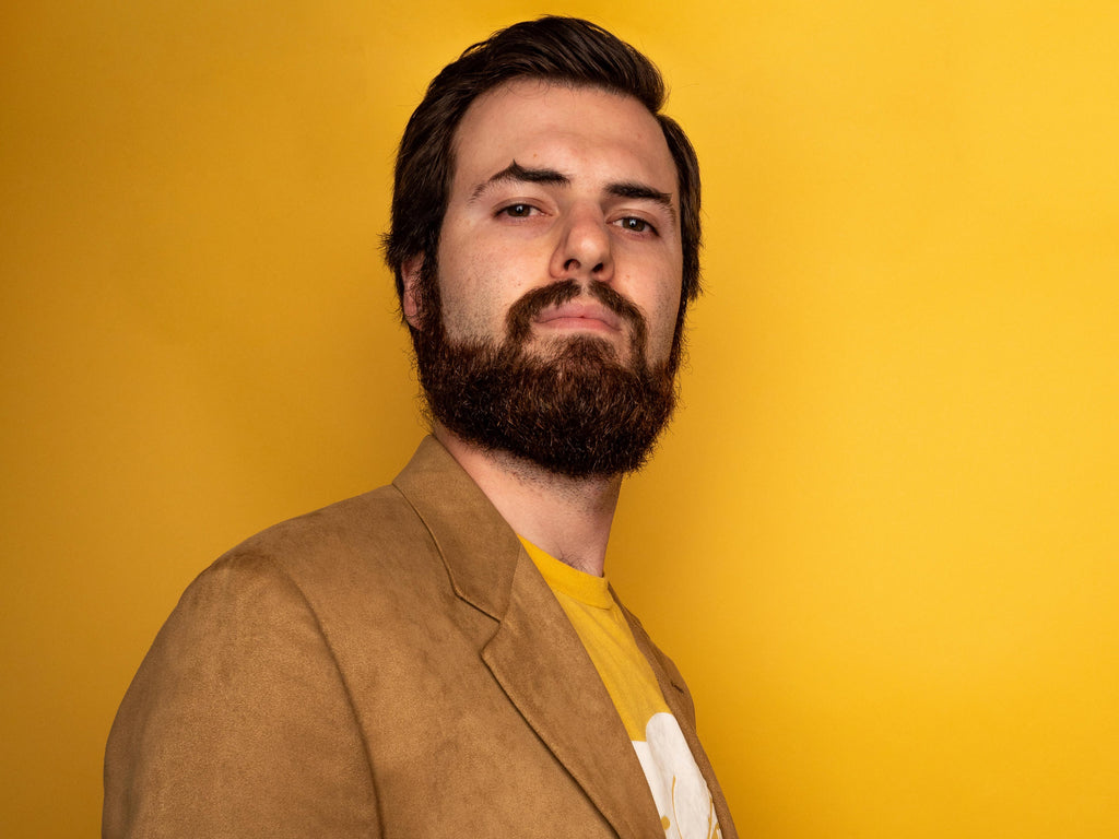 Brown haired man with a Hollywoodian beard posing in front of a yellow backdrop