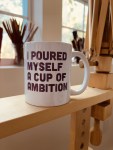 A white mug with bold black text that reads "I POURED MYSELF A CUP OF AMBITION" sits on a wooden surface, with blurred windows and brushes in the background.