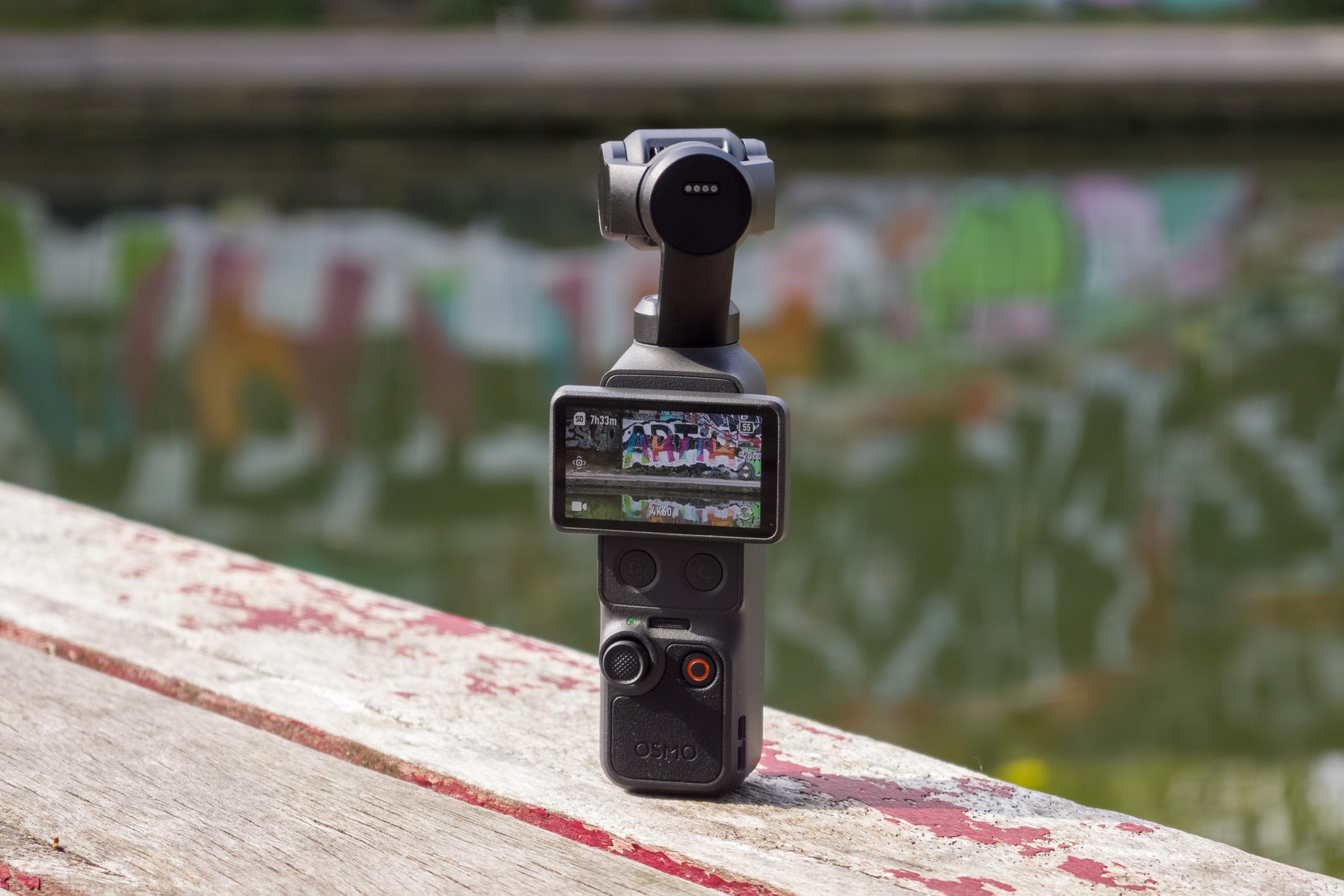 Photo of DJI Osmo Pocket 4 standing on a wooden table in front of a canal, showing the screen with the camera view.