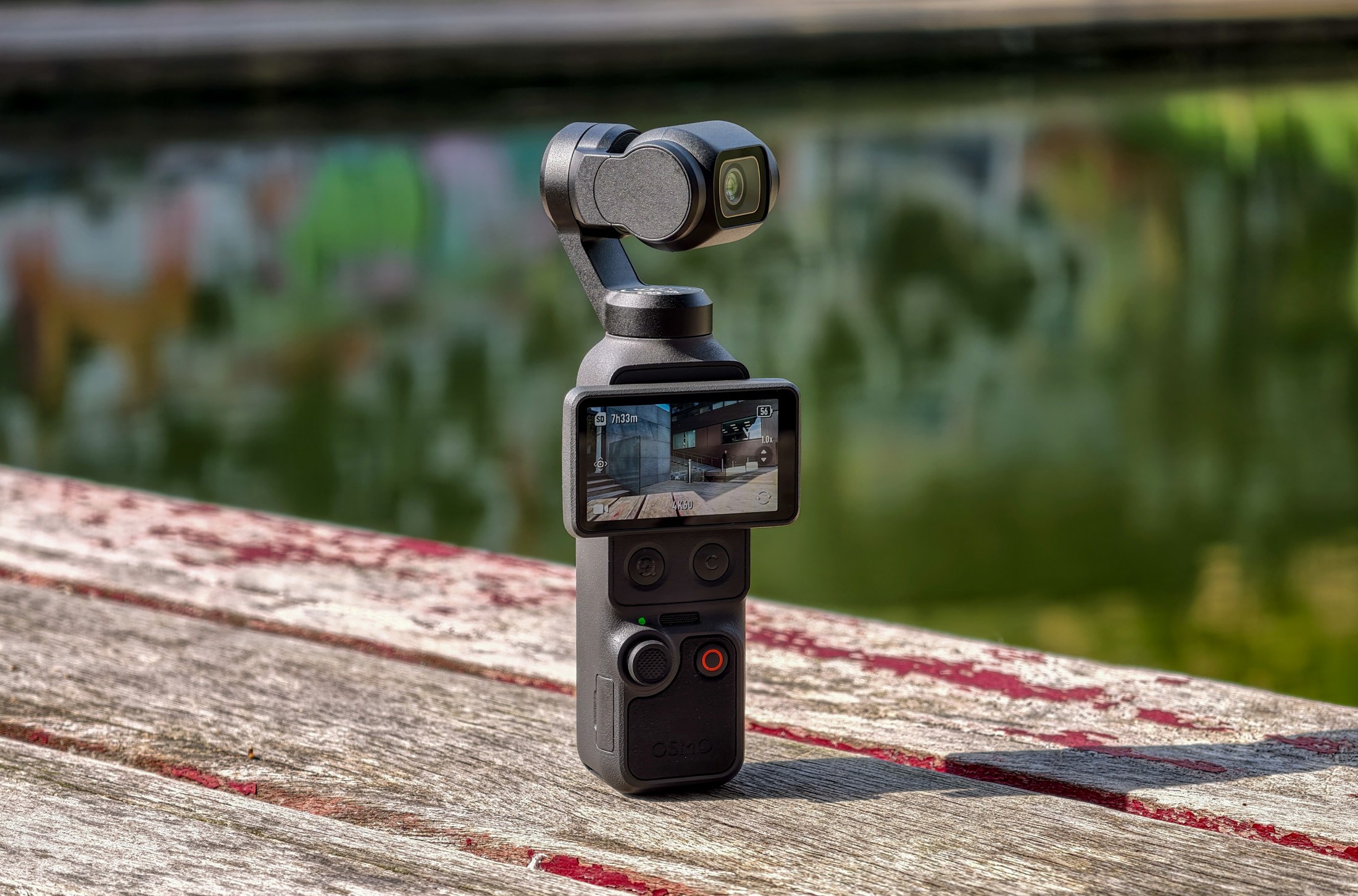 Photo of DJI Osmo Pocket 4 standing on a wooden table in front of a canal, showing the screen with the camera facing away