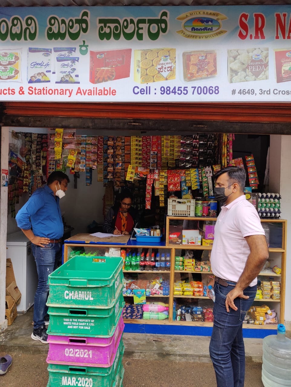 (L) Milan Partani, Business Head of Meesho SuperStore and (R) Dipak Nachani from Meesho's Business Finance team, talking to a retailer during a field visit in Mysore. Both men are brown, south-asian wearing shirts and jeans standing outside a small grocery shop selling every day groceries including dry groceries, biscuits, soaps, eggs, bottled water, etc.