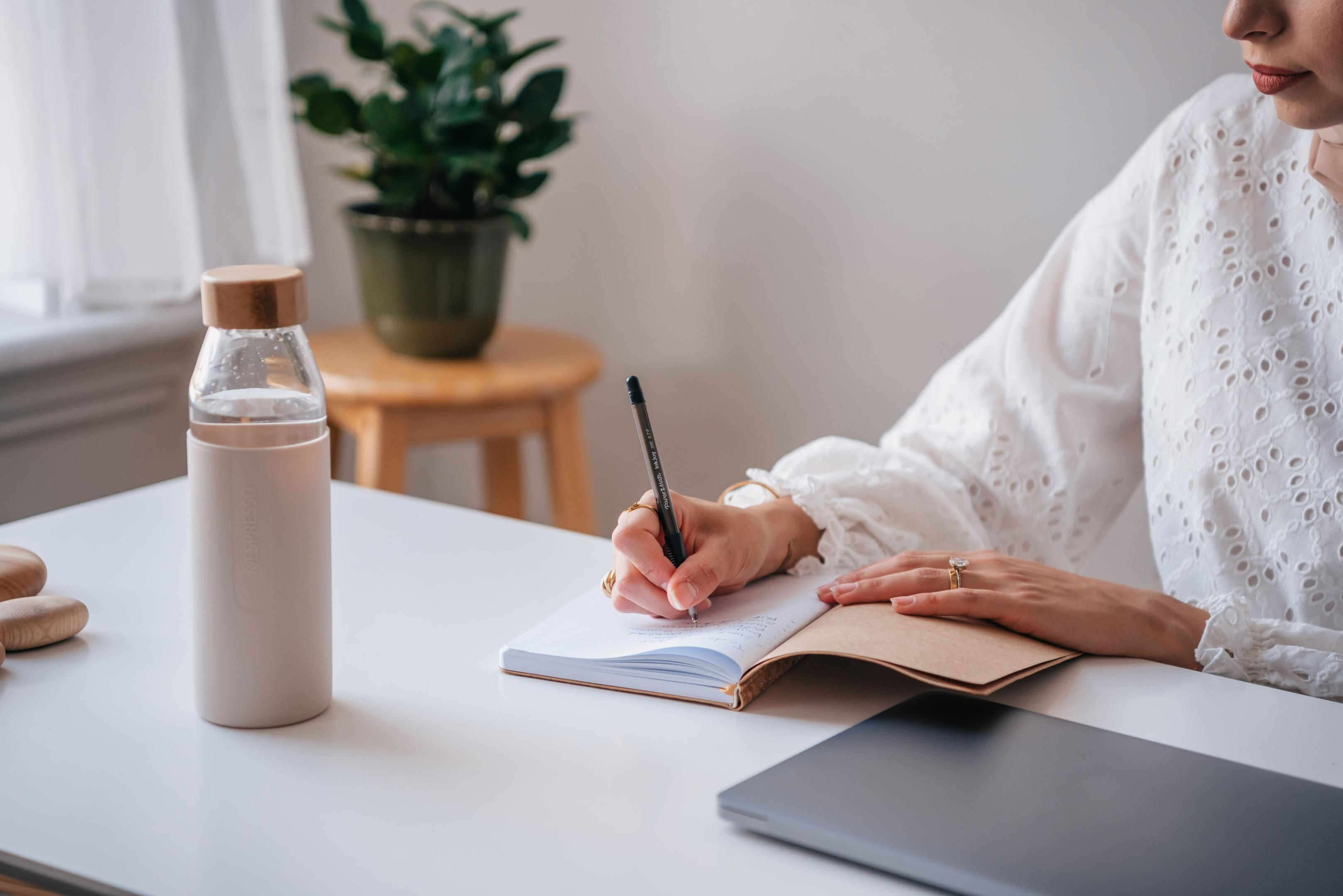 Woman at desk working writing in notebook with reusable water bottle by her.