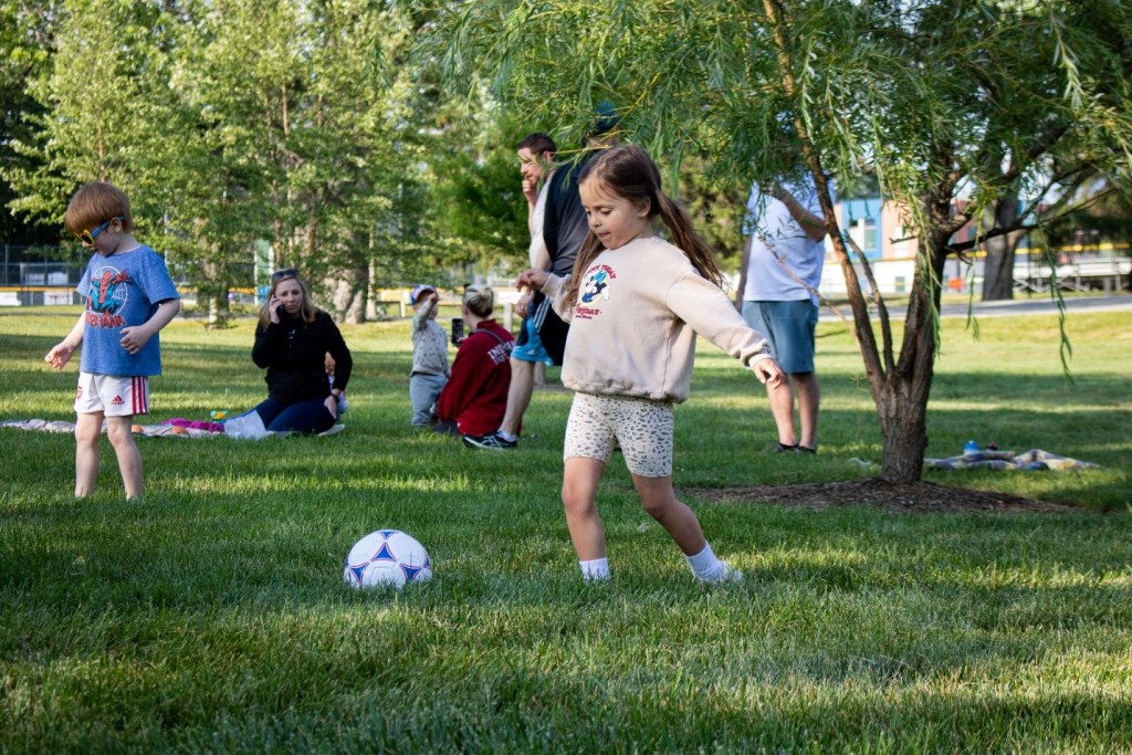 A young girl in a beige sweater plays with a soccer ball on a grassy area, surrounded by other children and adults in a park setting.