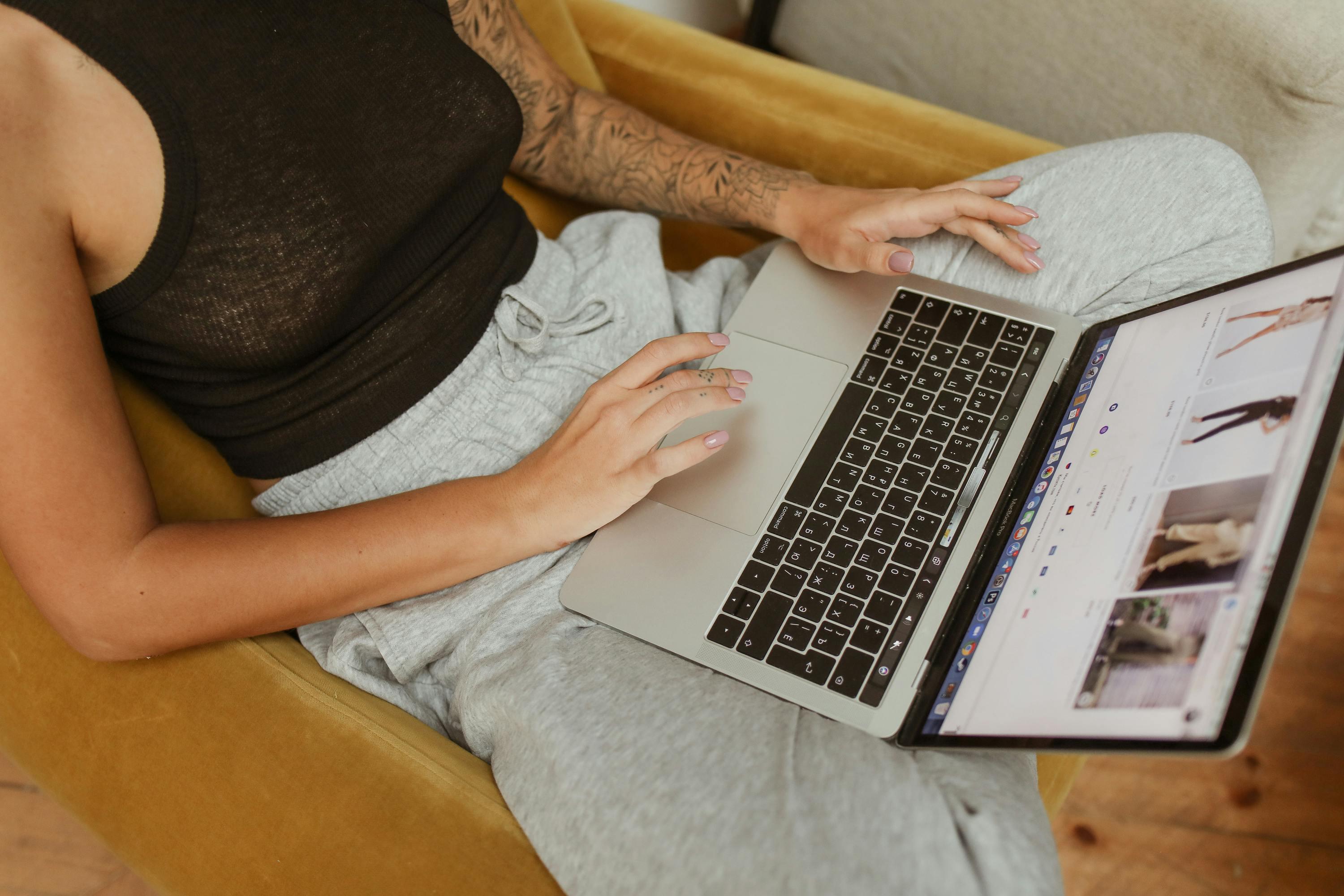 A woman is sat on a yellow chair scrolling through a website on a laptop. She is wearing grey sweatpants and a black tank top, with a floral pattern tattooed down her left arm.