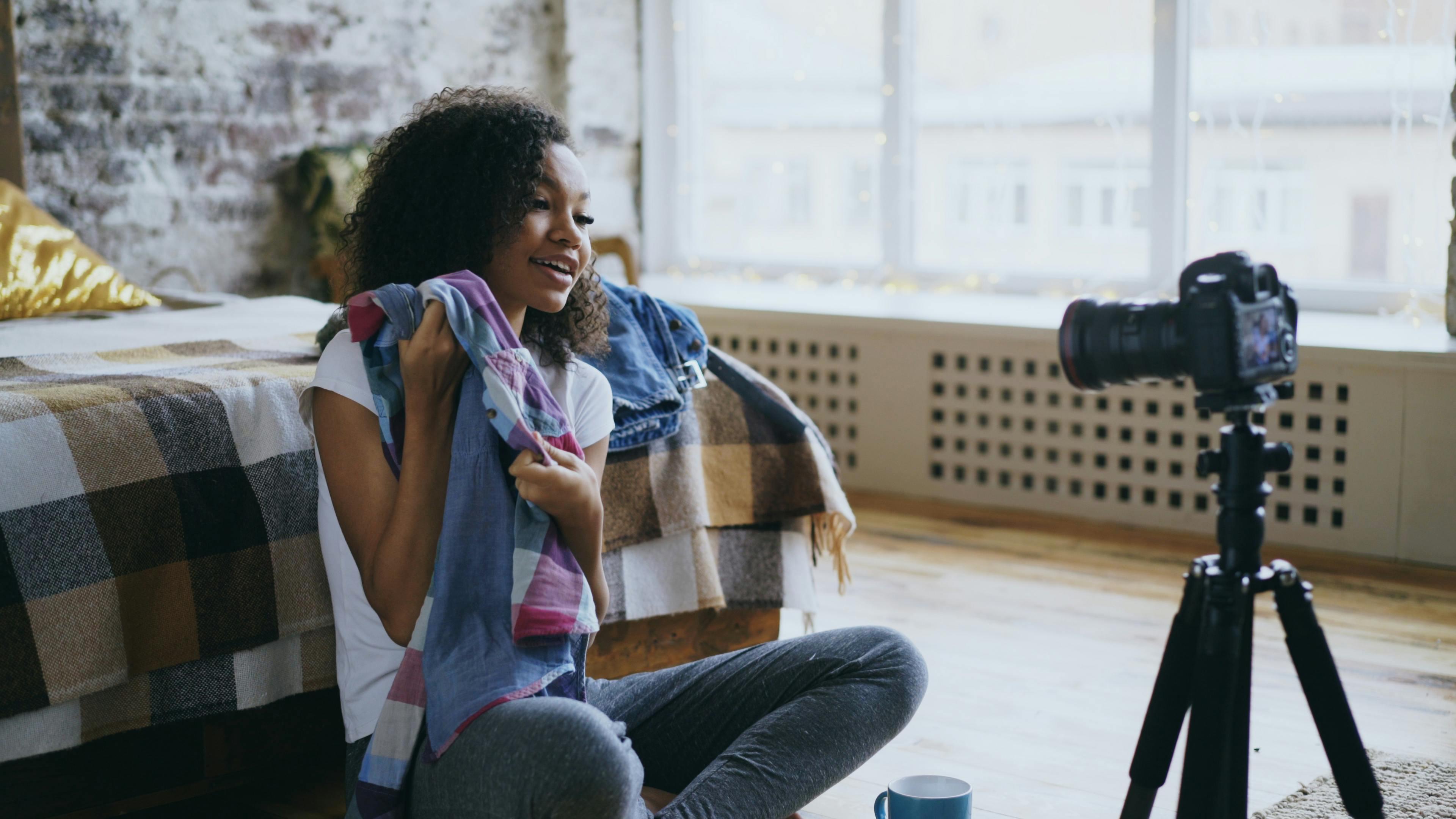 A young girl poses in front of a DSLR camera and tripod with items of clothing in her hands. 